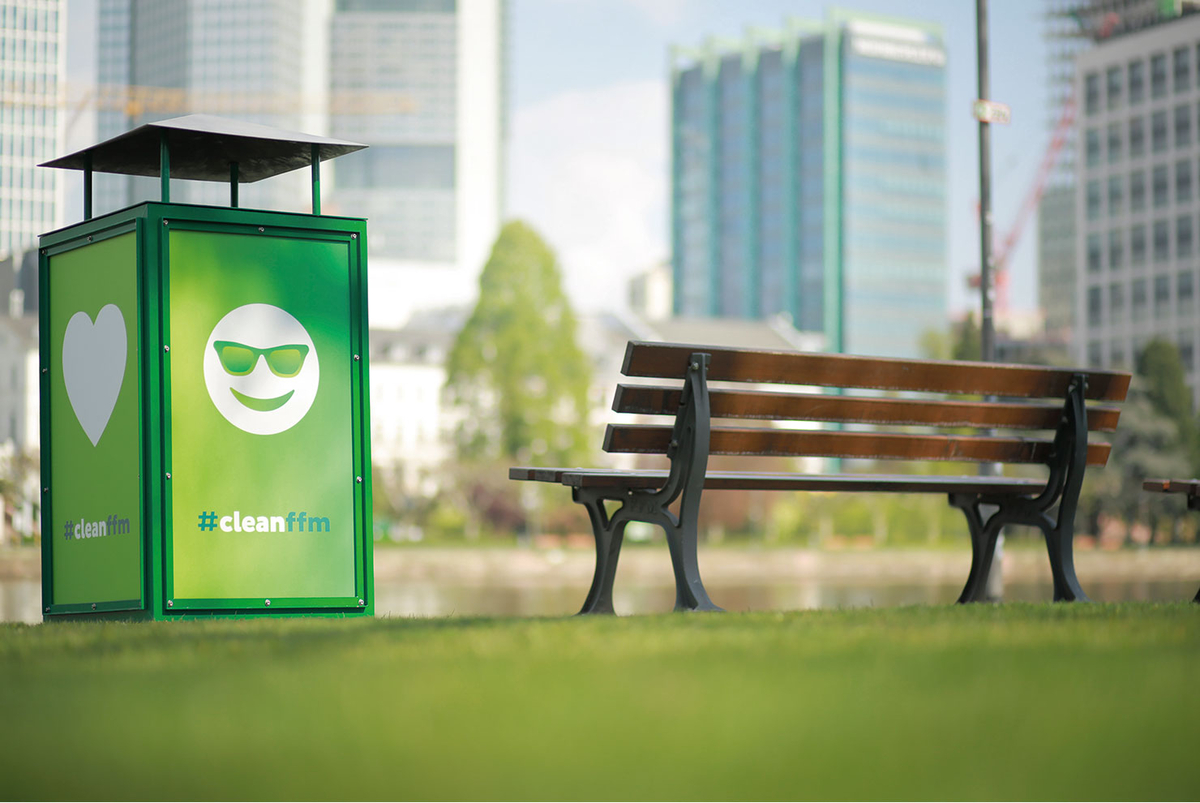Grüner Mülleimer mit cleanffm-Logo neben Parkbank im Stadtpark