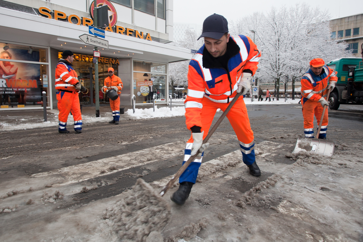 Mitarbeiter räumt Schnee auf Gehweg mit Schneeschieber