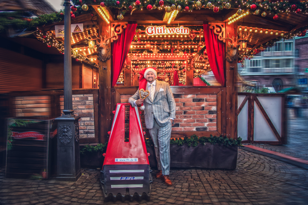 Kleider machen Leute 2026 - Mitarbeiter steht im Maßanzug auf dem Weihnachtsmarkt und lehnt sich an eine mobile Straßensperre