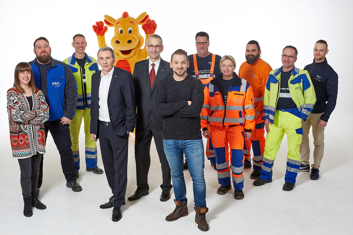 Mitarbeitende in bunter Arbeitskleidung stehen gemeinsam vor grauem Hintergrund